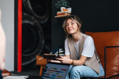 A woman sits on a large brown armchair, holding a tablet computer