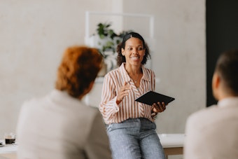 A woman smiles as she presents to two people, she is holding a tablet computer