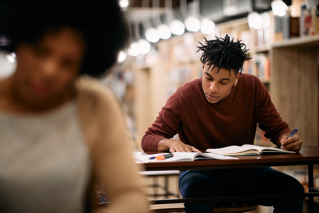 A young male student in a brown jumper sits at a desk with a pen and notepad