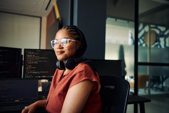 A woman with headphones sits at a desk, screens with code are behind her