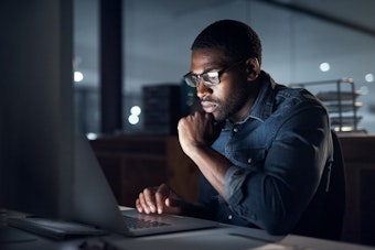 A man with glasses sits in an office in the evening, looking at laptop computer