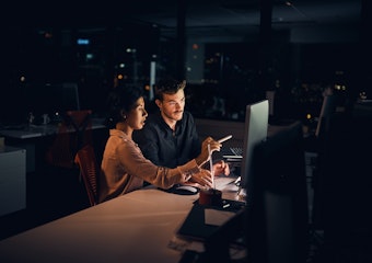 A man and woman look at a desktop computer in a dark office room