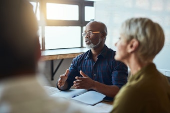 A man in a blue shirt and glasses speaks to across a table to a group of people