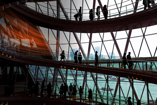 A multi-story glass building with people walking across walkways. A city can be seen in the background through the windows.