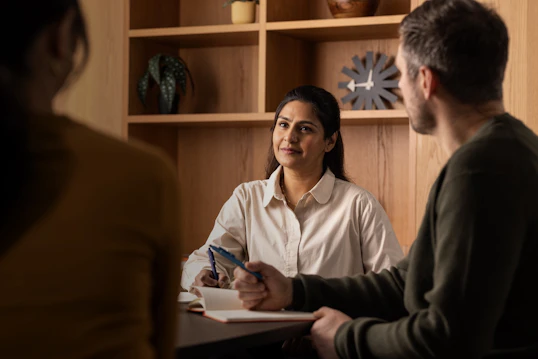 Men and women with notebooks have a discussion across a desk. They are in a modern, wood panelled office with shelves adorned with clocks and plants