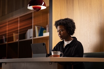A woman sits at a desk in a modern office with a tablet computer and tea
