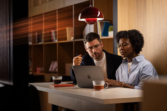 A man and woman sit in a coffee shop. They are looking at a laptop computer.