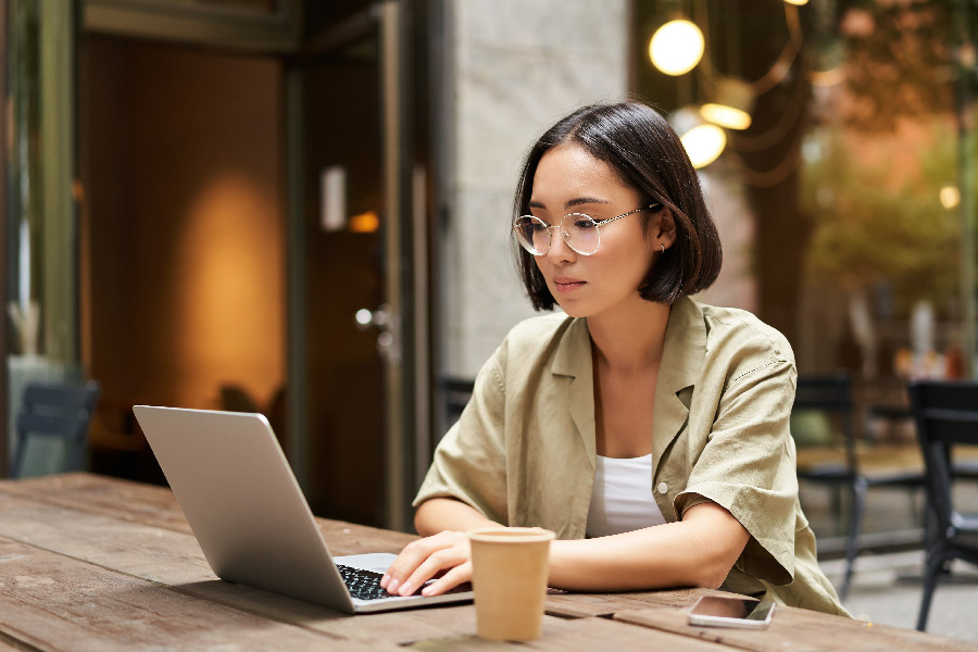 A woman with glasses sits outside with a laptop and coffee at a wooden bench
