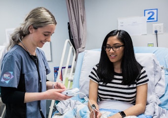 A female nurse smiles as she uses a machine to check the heart rate of a patient lying in a blue hospital bed