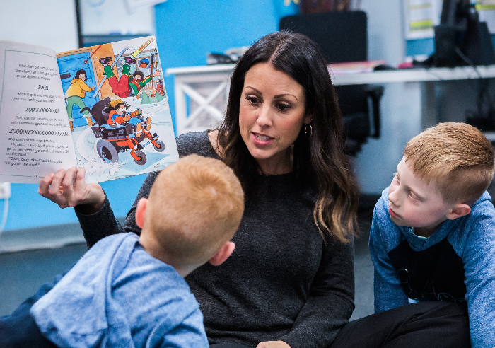 A female nursery teacher holds up a picture book as she reads to two small children