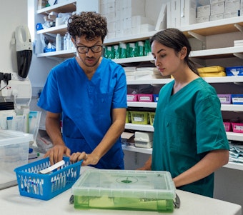 A male and female pharmacist in inspect a series of containers containing different form of medication. They are wearing scrubs and are standing infront shelves of medical supplies