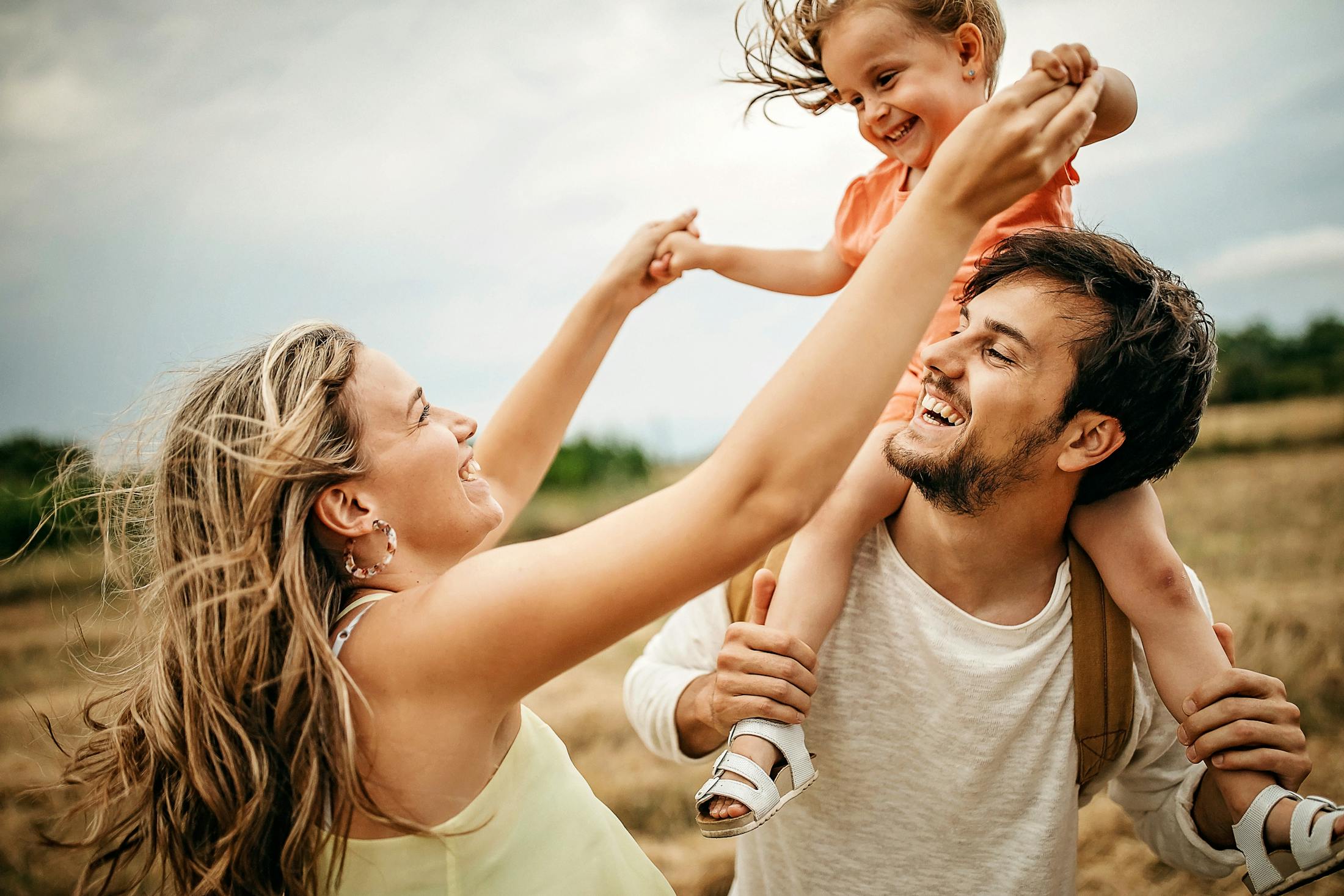 Dad and mom playing with their daughter in a field. The daughter is about 3 years old and on her dads shoulders