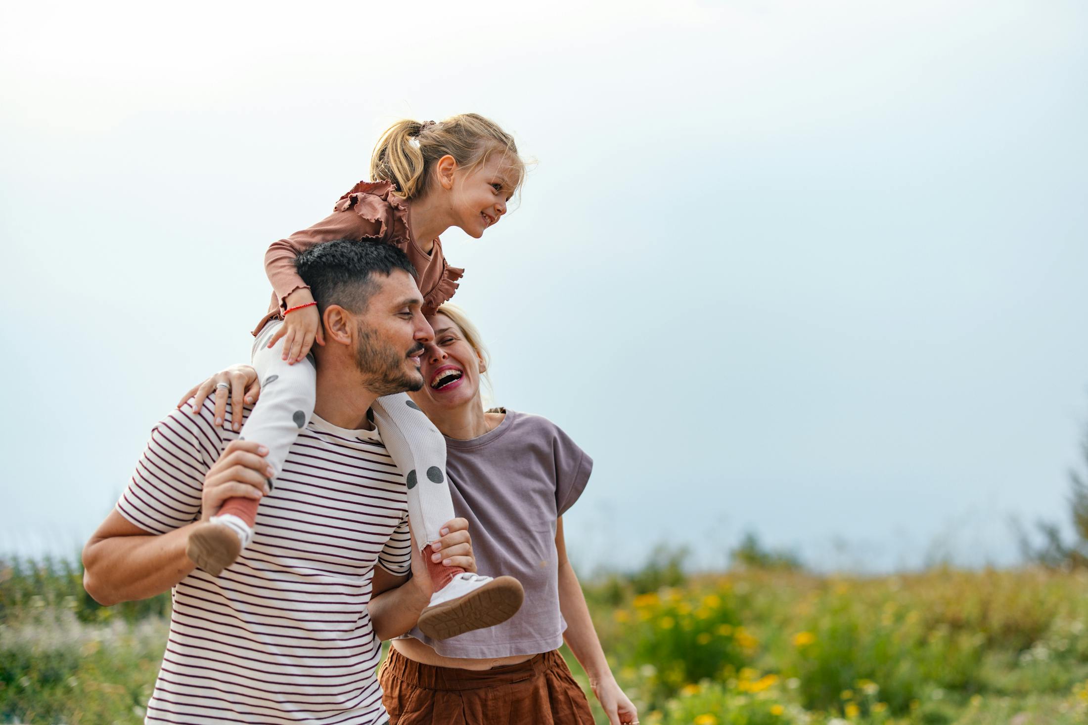 happy family going on a walk next to a meadow. A little firl sits on her father's sholders