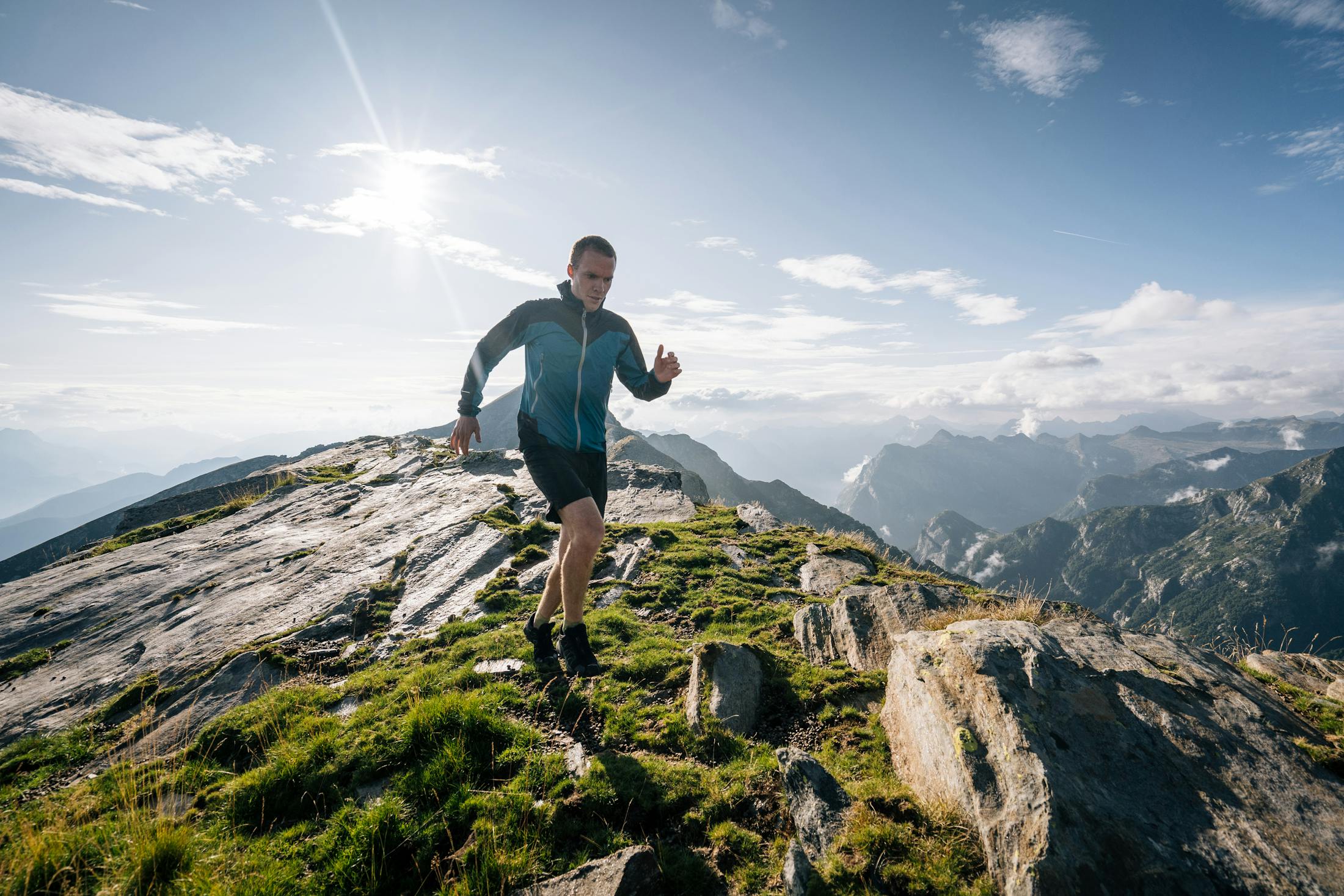 man running on a very high mountain top surrounded by other mountains with a blue sunny sky.