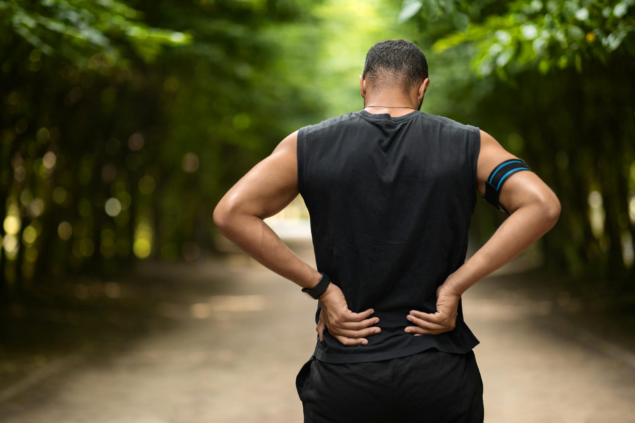 Man holding his back in pain after a run through a forest lined park.