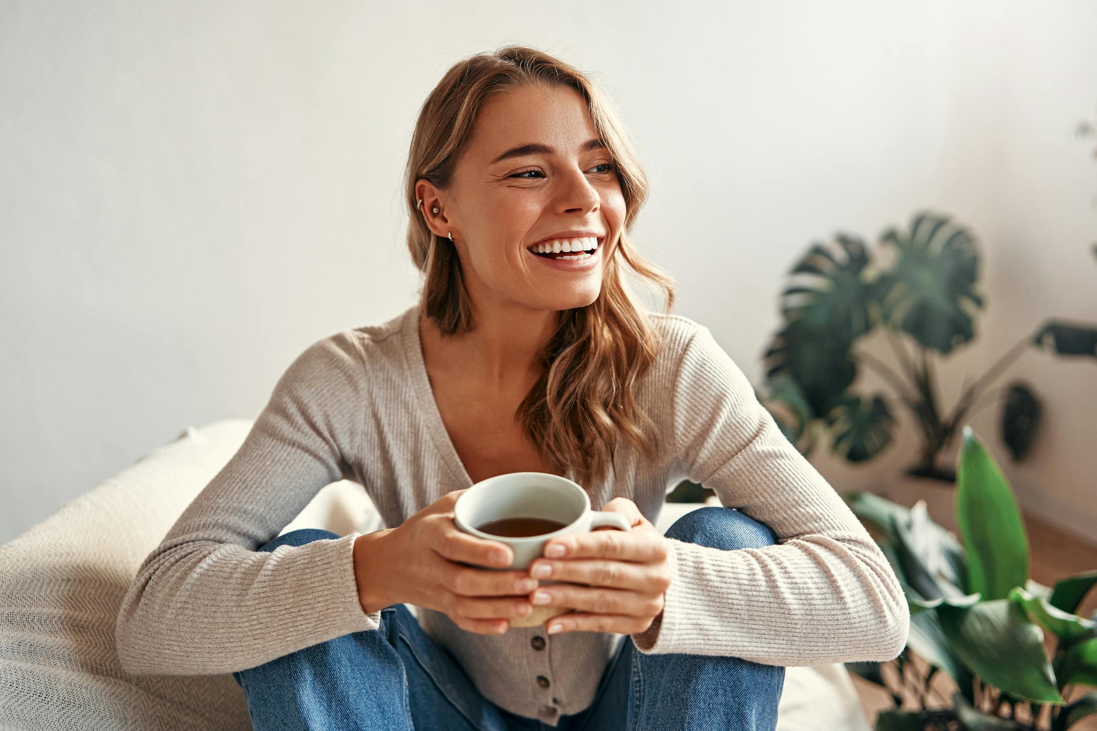 Woman is smiling while sitting on her couch drinking a cup of coffee