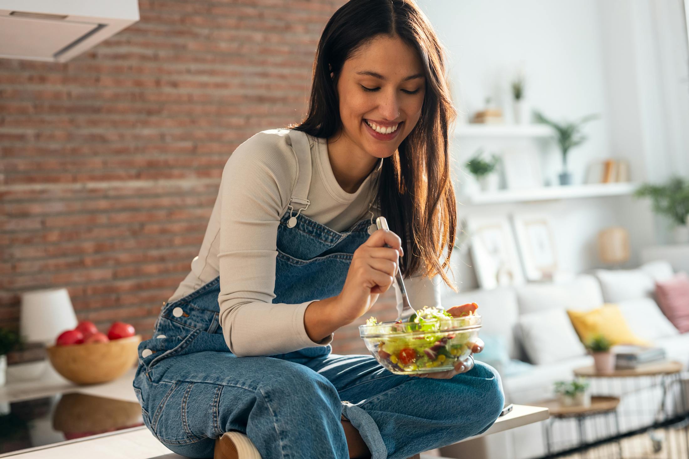 Woman siting on her kitchen counter smiling as she eats a salad.