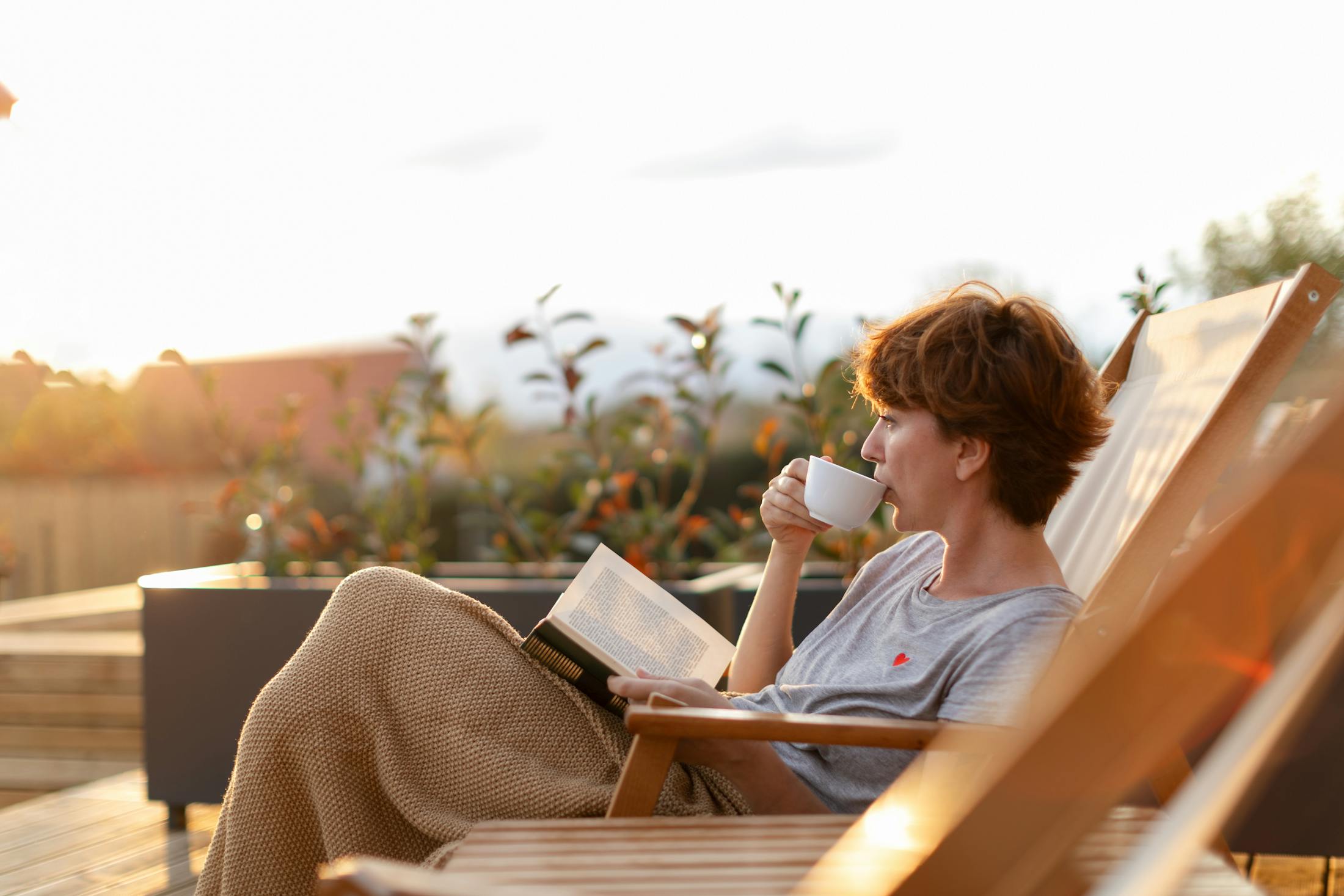 Woman drinking coffee and reading a book with a blanket on her lap as she lounges in a chair outside