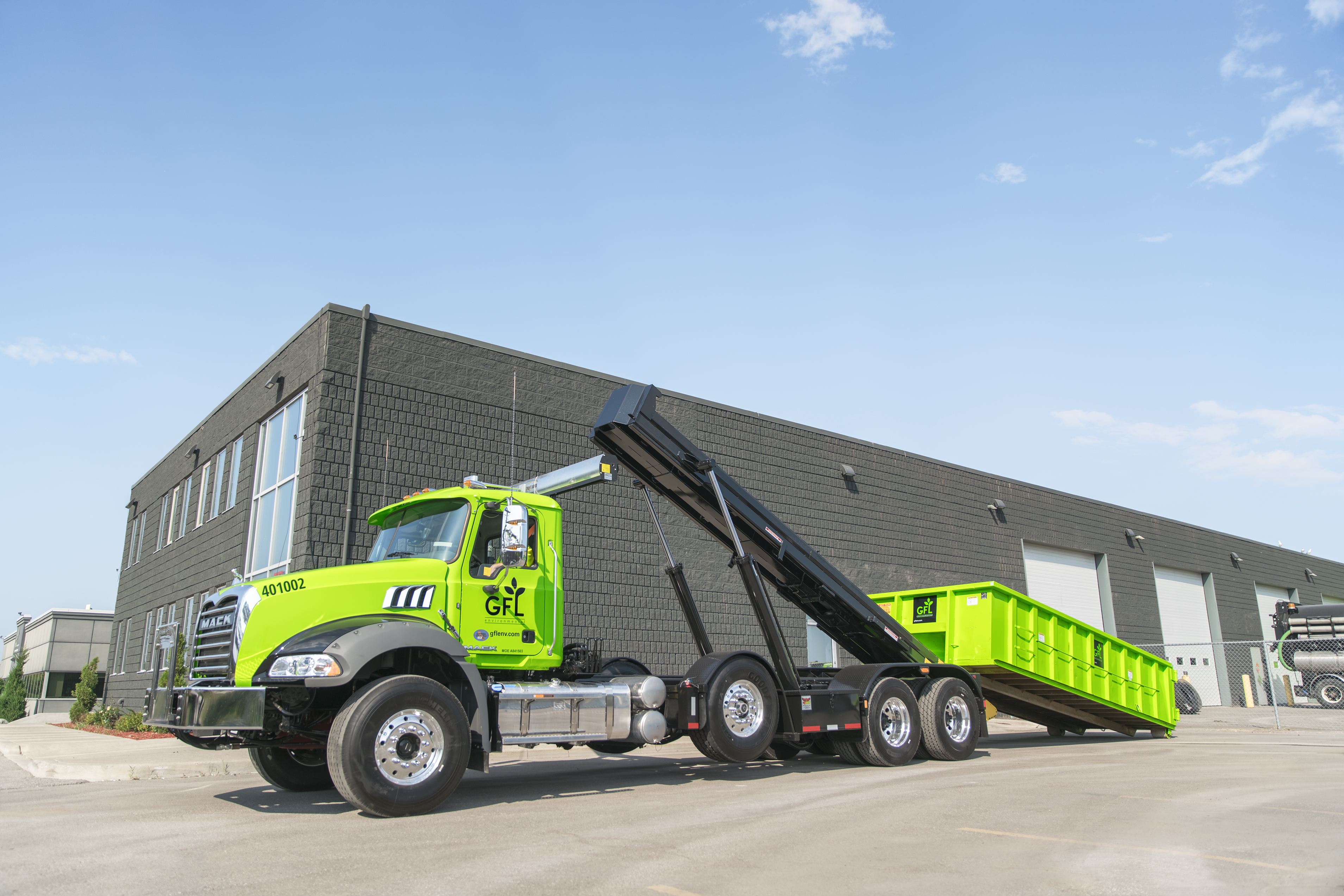 A bright green GFL roll-off truck with an elevated bin parked in a facility yard in front of a brick building