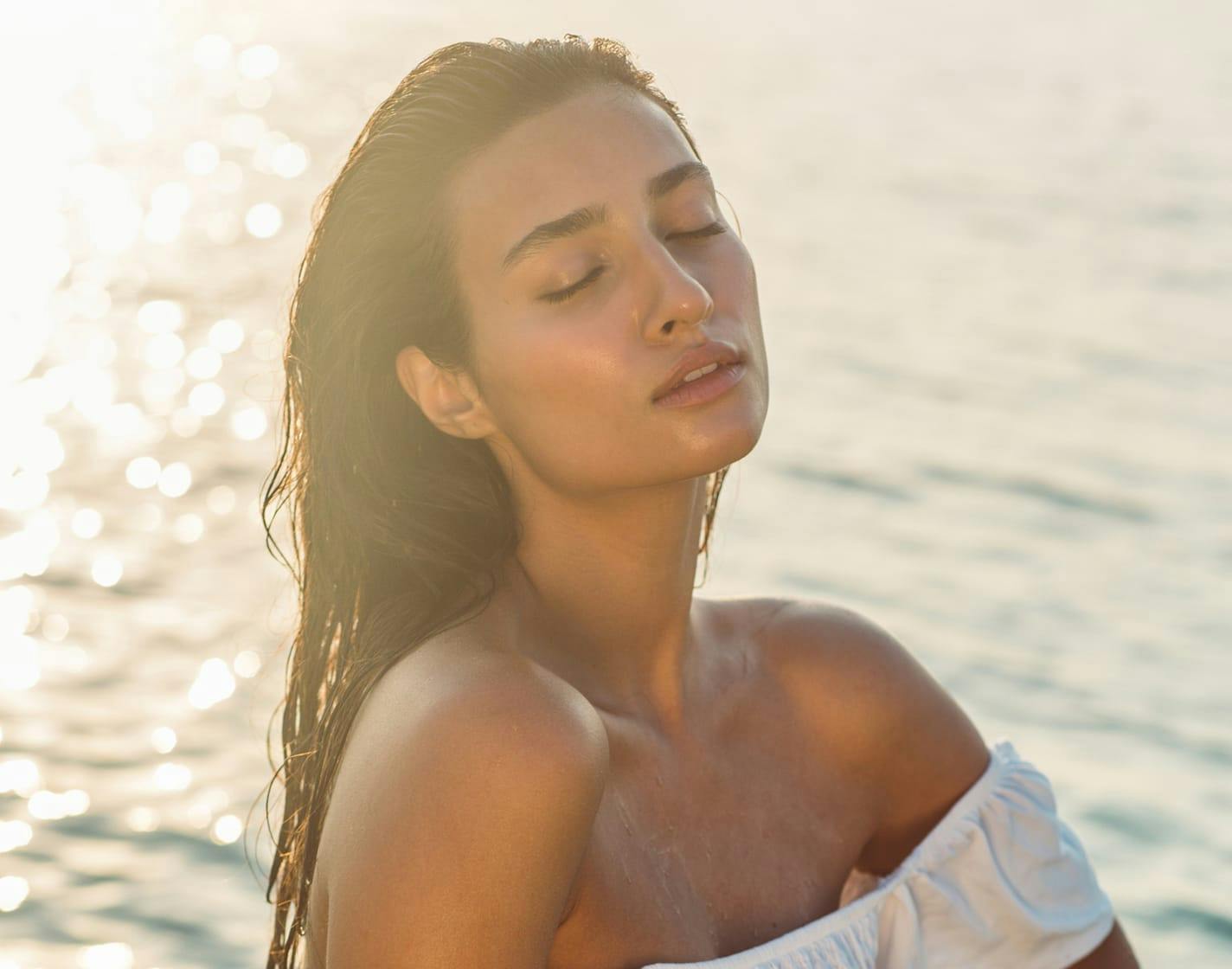 Dark haired woman sitting next to water in a white off the shoulder top