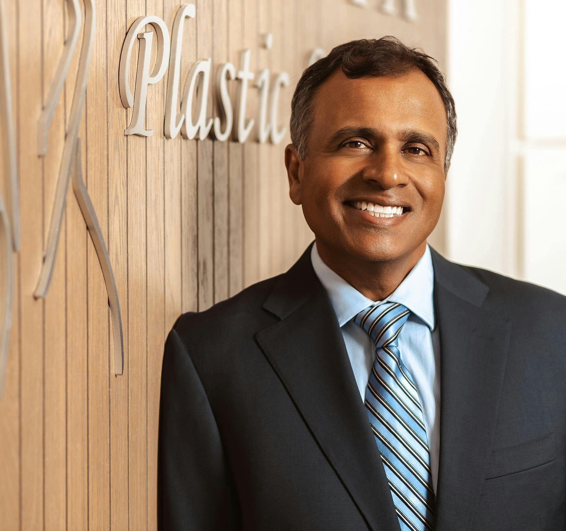 Dr. Gupta standing in front of Gupta Plastic Surgery logo in office