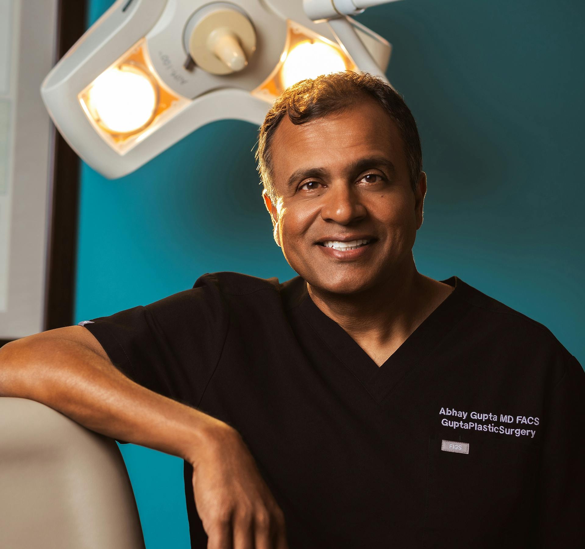 Dr. Gupta in black scrubs leaned against medical chair in Gupta Plastic Surgery office
