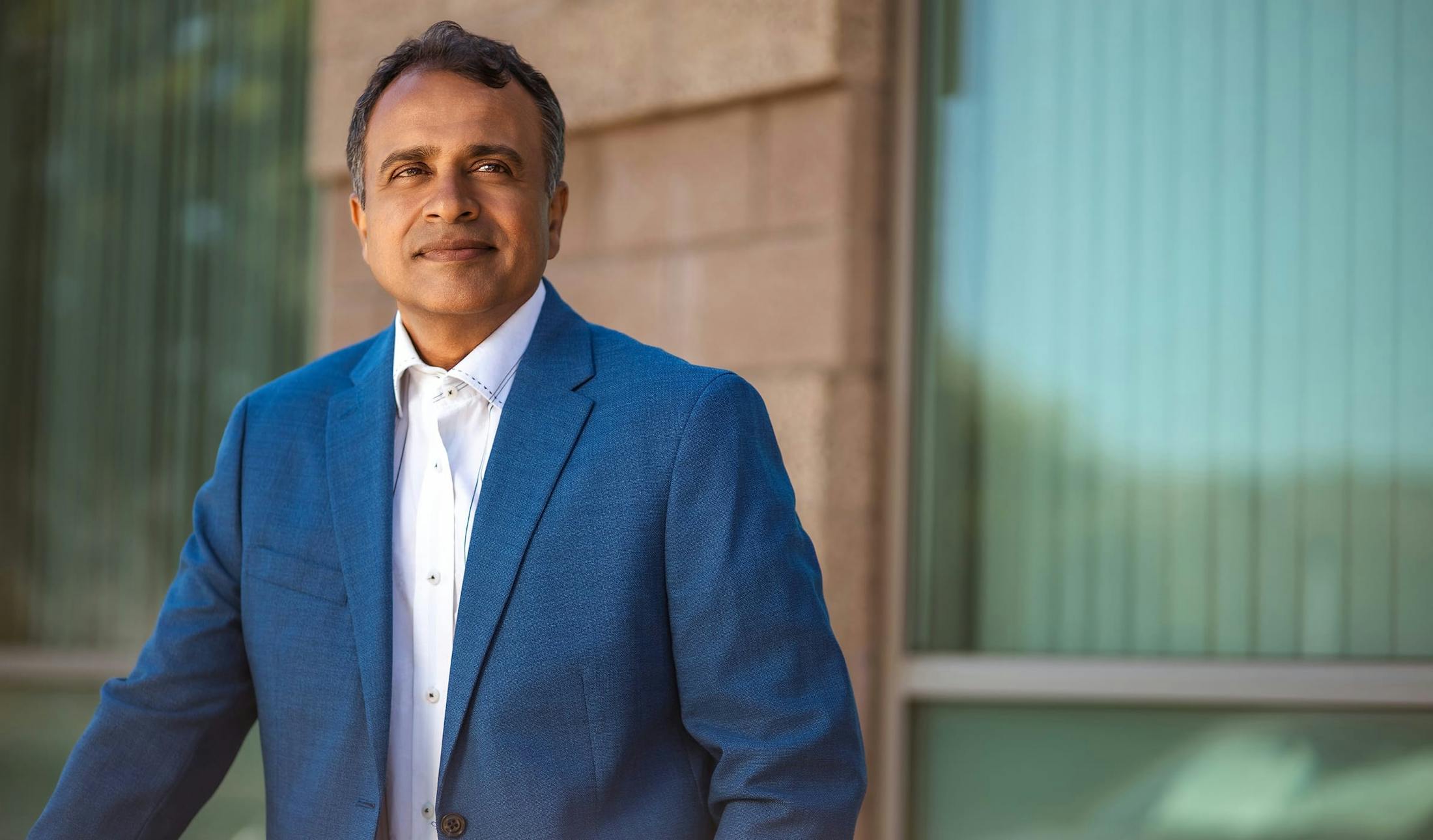 Dr. Gupta in navy suit jacket and white button up shirt standing in front of windows