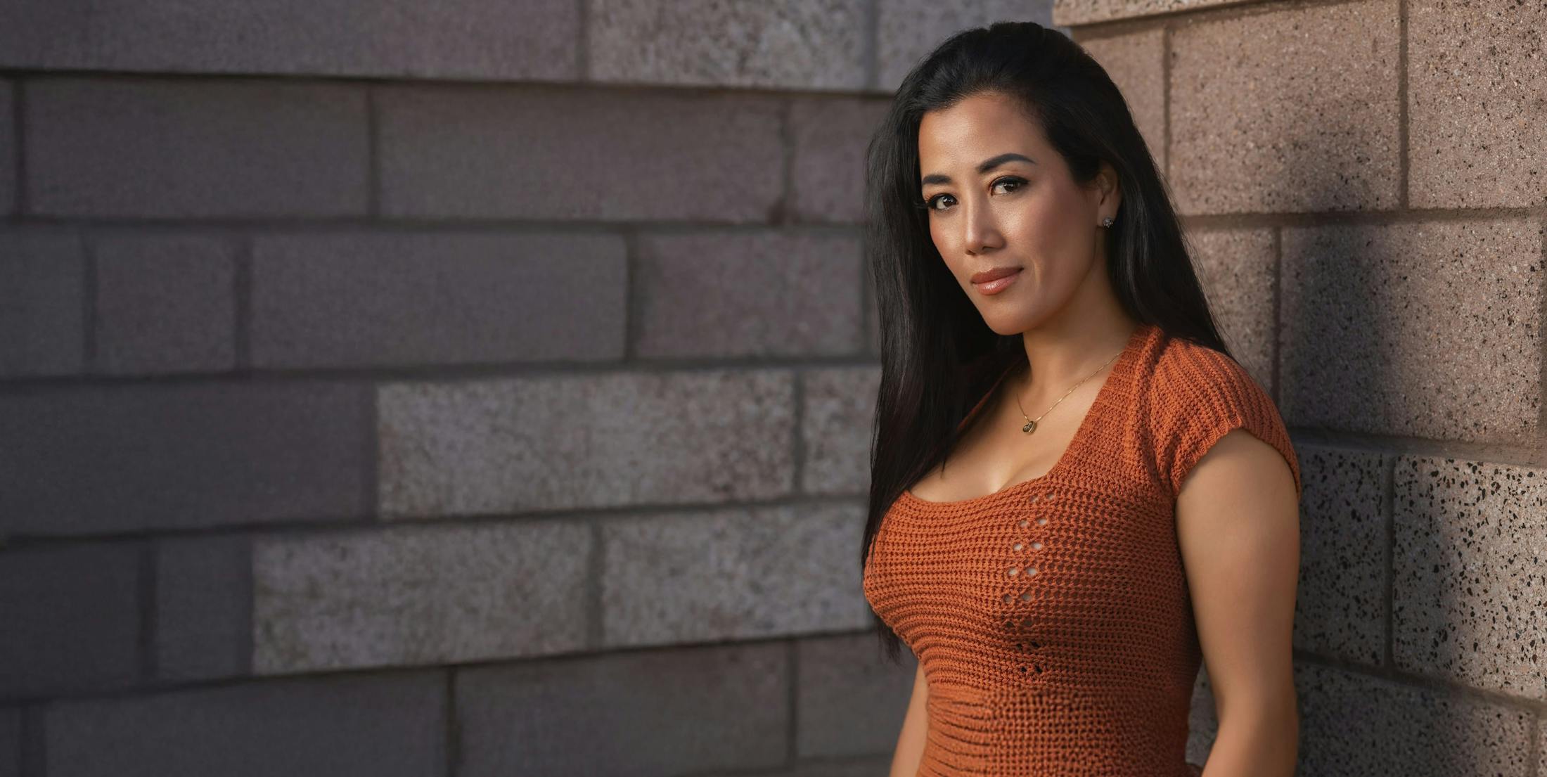 Woman in orange shirt with long black hair standing agianst brick wall