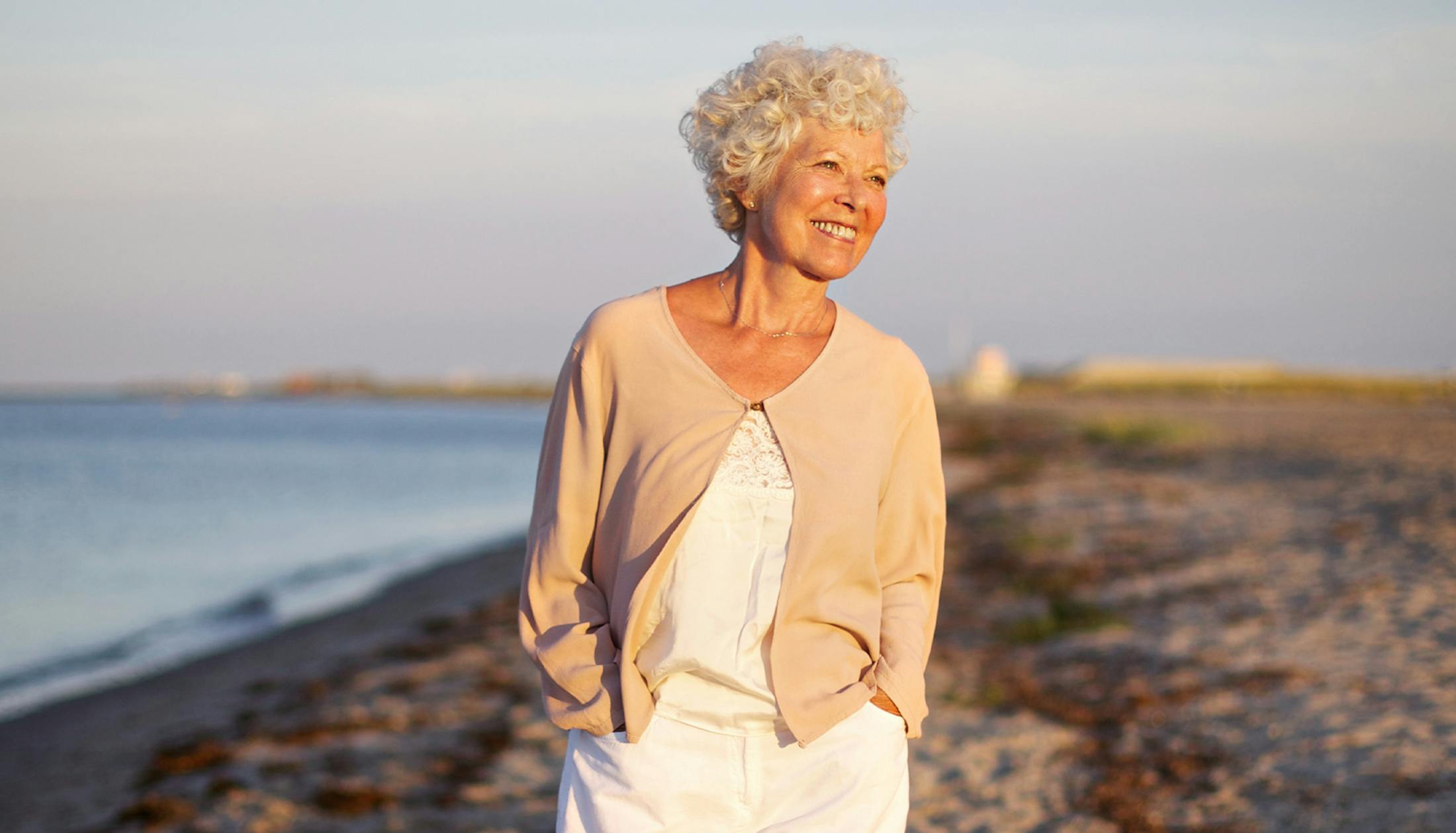 older woman walking on the beach with her hands in her pockets