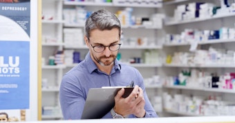 A male pharmacist wearing glasses stands in a dispensary holding a clipboard