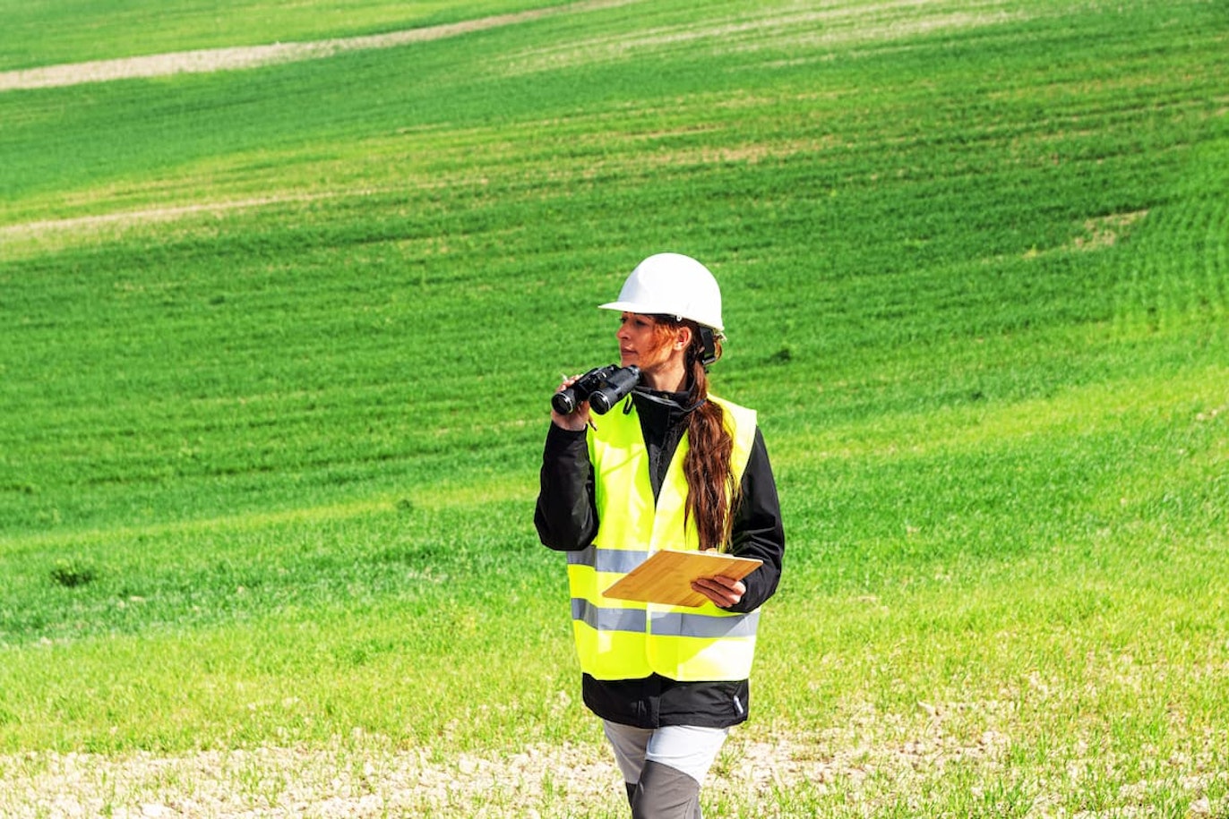 Female engineer inspecting green field with binoculars and clipboard