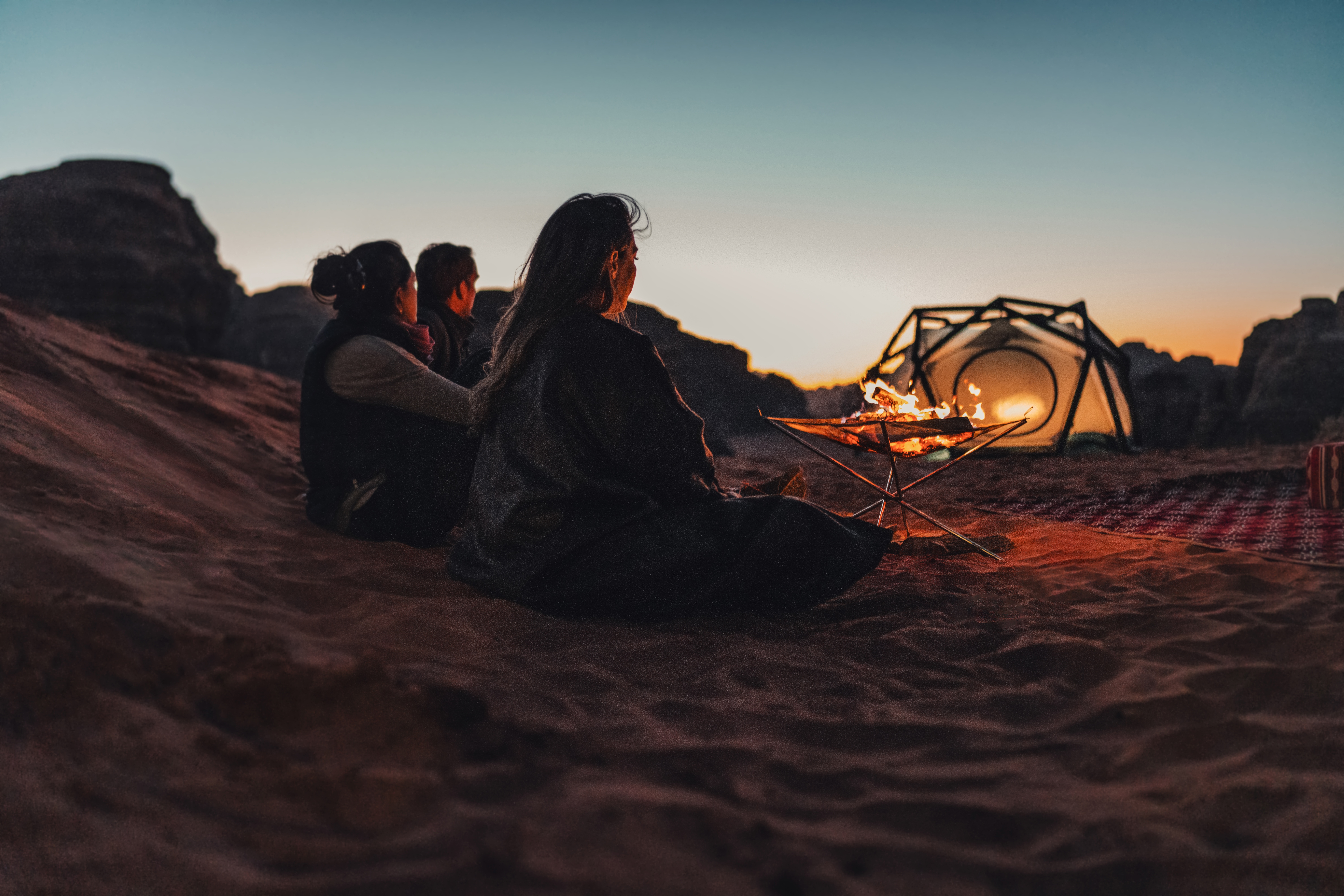 Three people sitting on sand, watching a fire and sunset in a desert landscape.