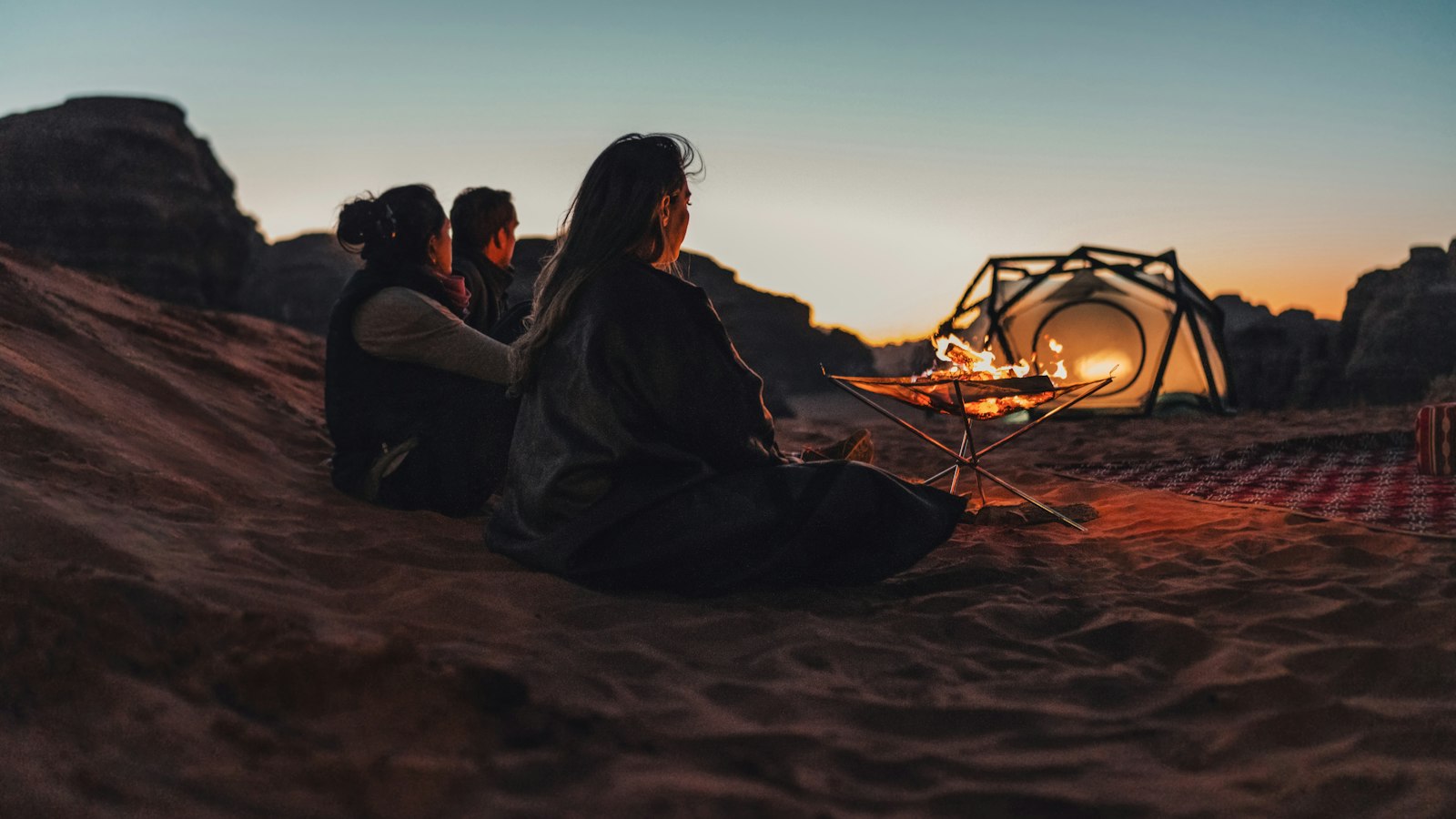 Another image Three people sitting on sand, watching a fire and sunset in a desert landscape.
