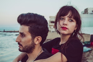 A man and a woman standing closely on a beach, with the woman slightly behind the man, gazing thoughtfully in different directions. The beach and ocean can be seen in the background during what appears to be sunset or dusk.