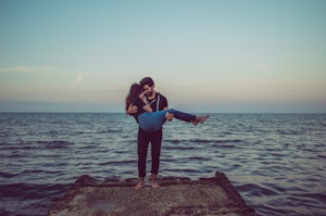 A person is standing barefoot on a concrete platform by the sea, holding another person in their arms. The sky is clear with a hint of clouds, and the water appears calm, reflecting shades of blue and gray as the sun sets on the horizon.