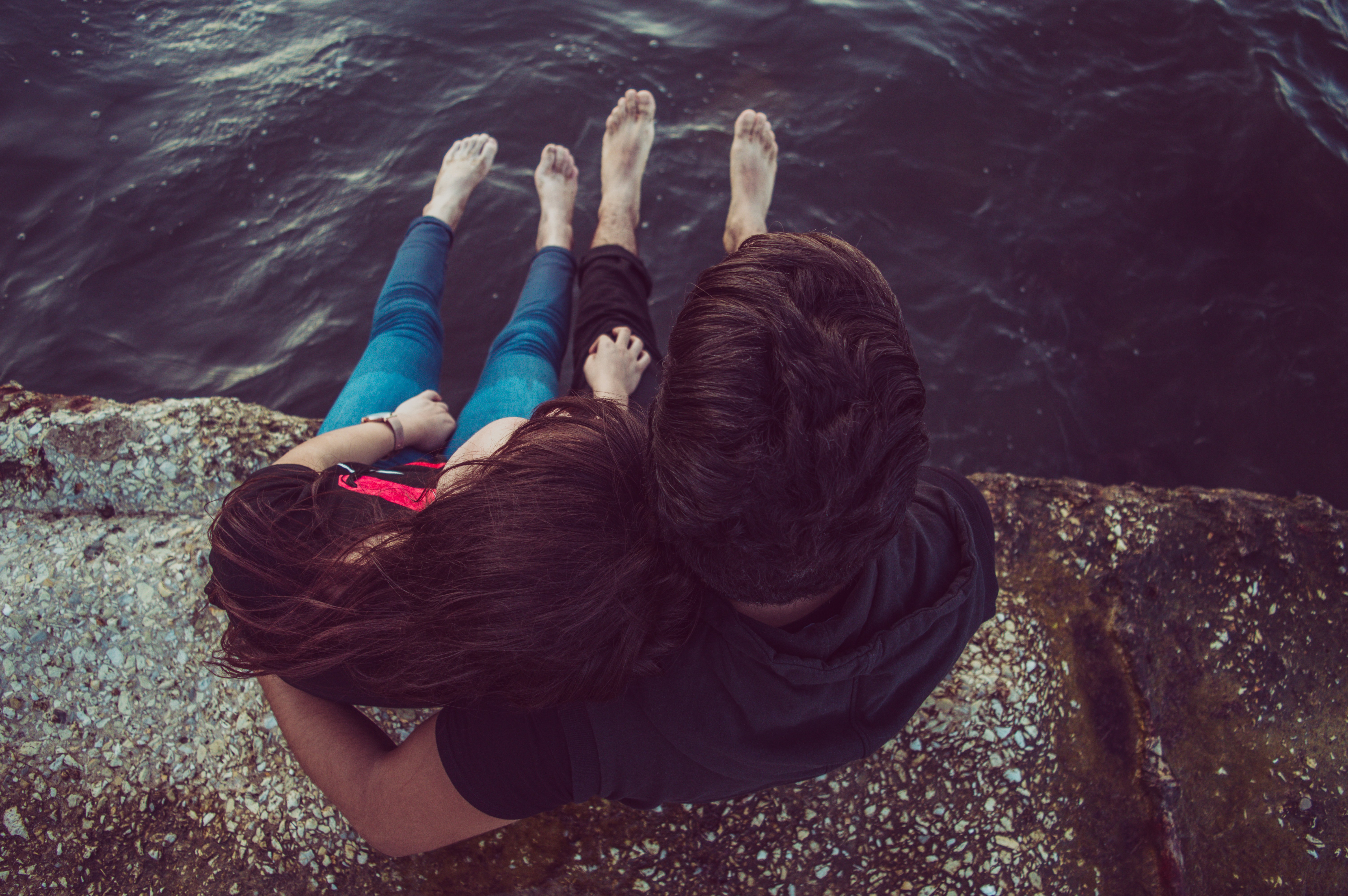 A couple is sitting on the edge of a rocky surface near a body of water. They have their feet dangling over the water, with one person resting their head on the other's shoulder. They are seen from above, creating a peaceful, intimate scene.