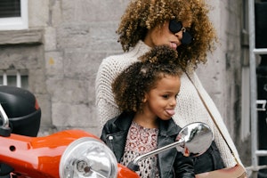 A woman in sunglasses and a cozy sweater poses playfully with a child on a red scooter, both sticking their tongues out, conveying joy and fun.