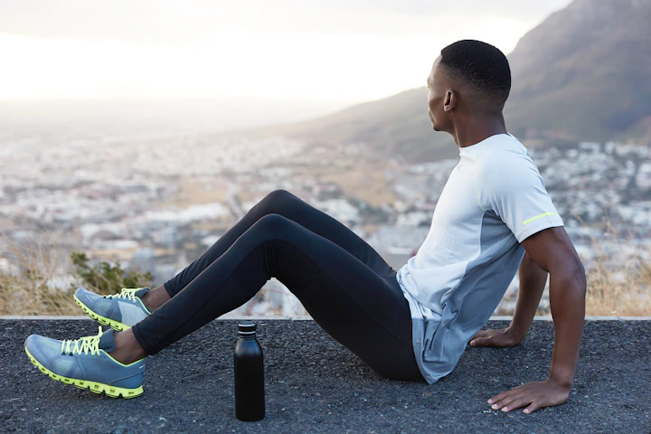 photo relaxed man with short hair dark skin sits highway with bottle cold water wears tracksuit focused aside enjoys spare time mountain view rests after workout fitness exercising 1
