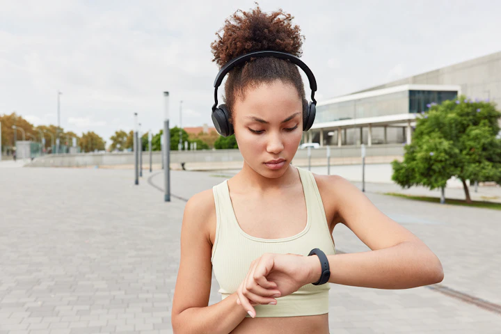 waist up shot sporty woman with combed curly hair uses smartwatch checks burned calories listens music via headphones wears cropped top poses outdoors stadium engaged active sports activity 1