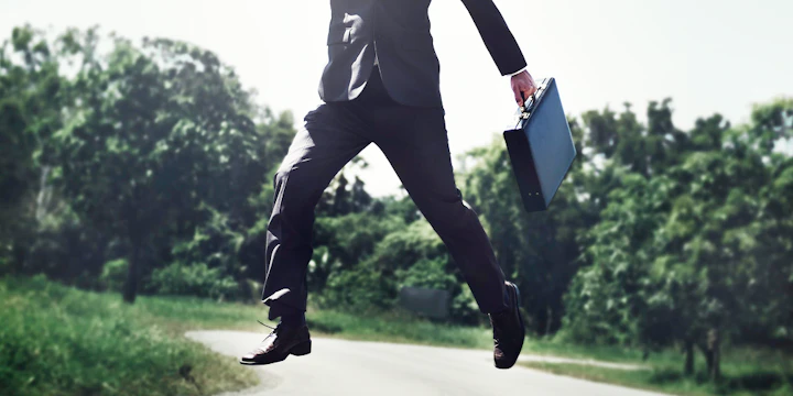 homme en costume noir avec un sac saute avec des arbres en arrière plan
