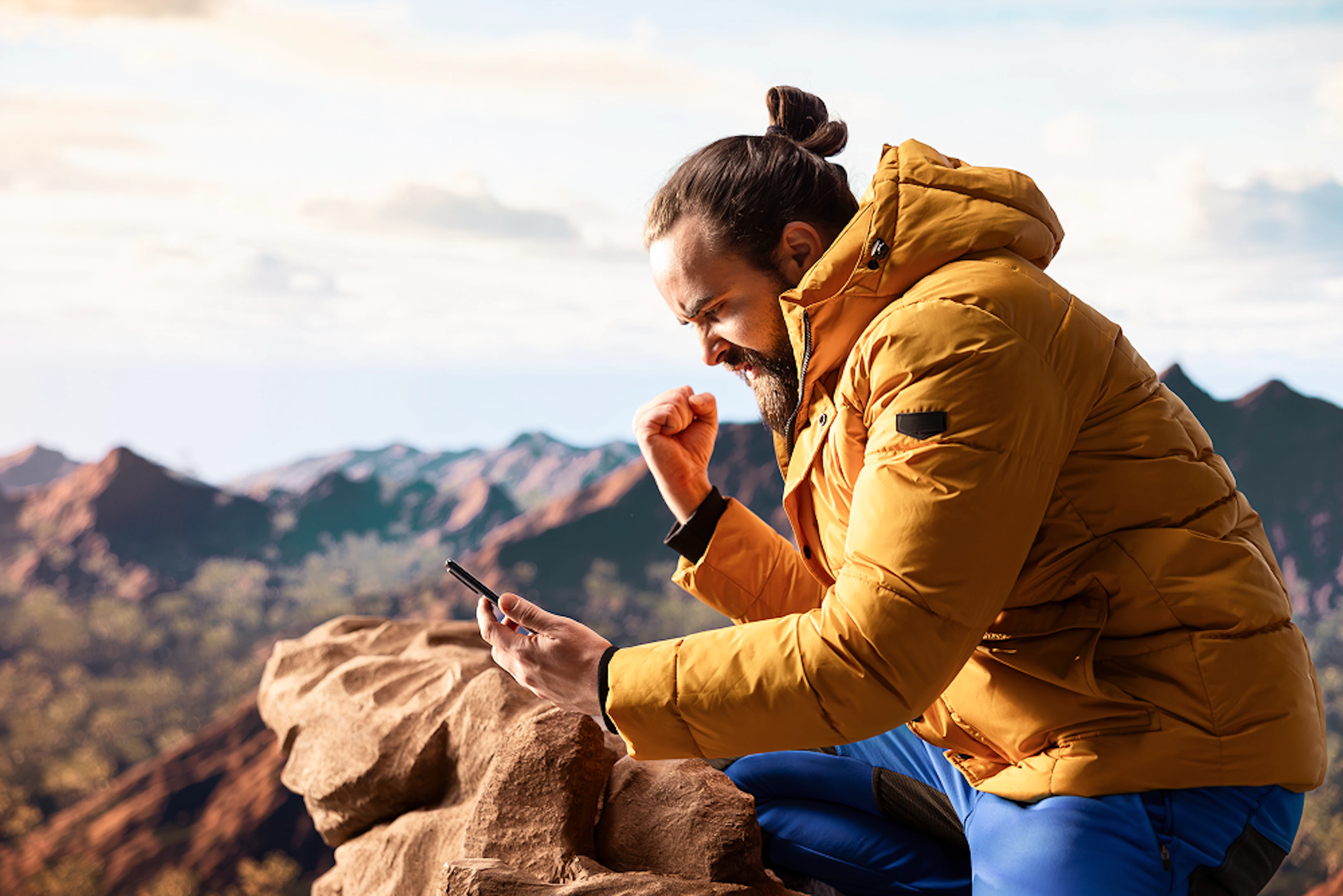 homme en doudoune jaune en montagne regarde son téléphone en brandissant le poing