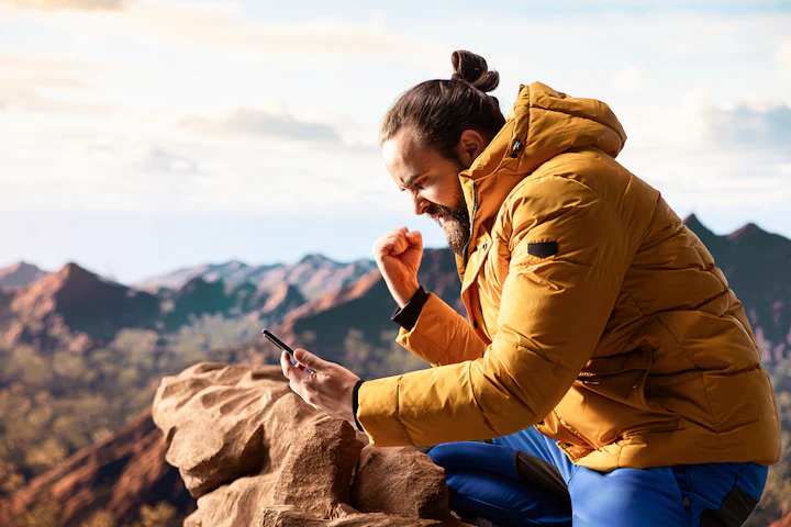 homme en doudoune jaune en montagne regarde son téléphone en brandissant le poing