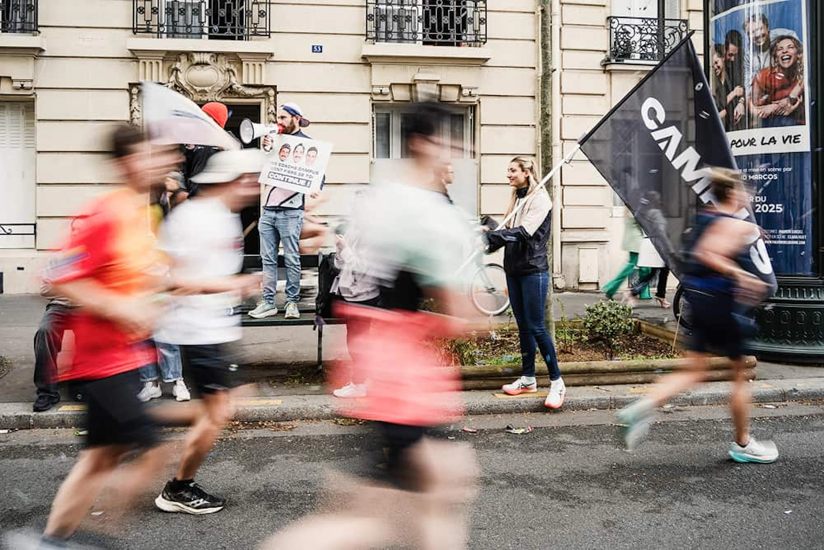 des coureurs flous courent devant des personnes qui tiennent un haut parleur et un drapeau Campus