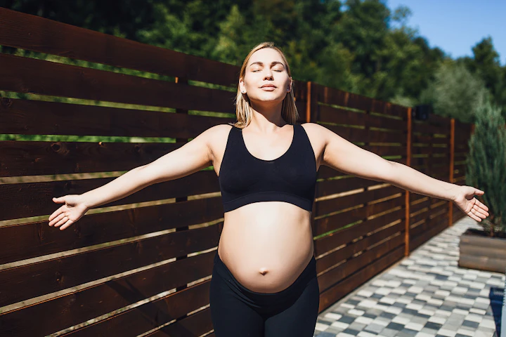 Une femme enceinte en tenue de sport ouvre les bras, le visage au soleil