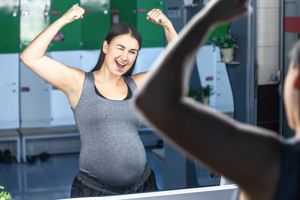 Une femme enceinte en tenue de course à pied lève les bras et fait un clin d'oeil face à un miroir