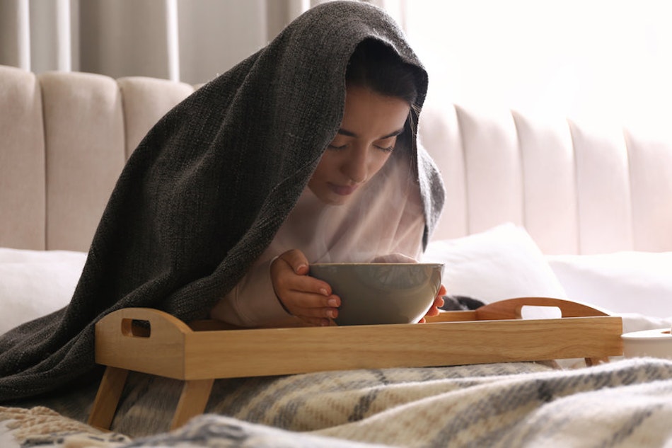 Person sitzt mit einer Schüssel für eine Dampfinhalation unter einer Decke auf einem Bett.