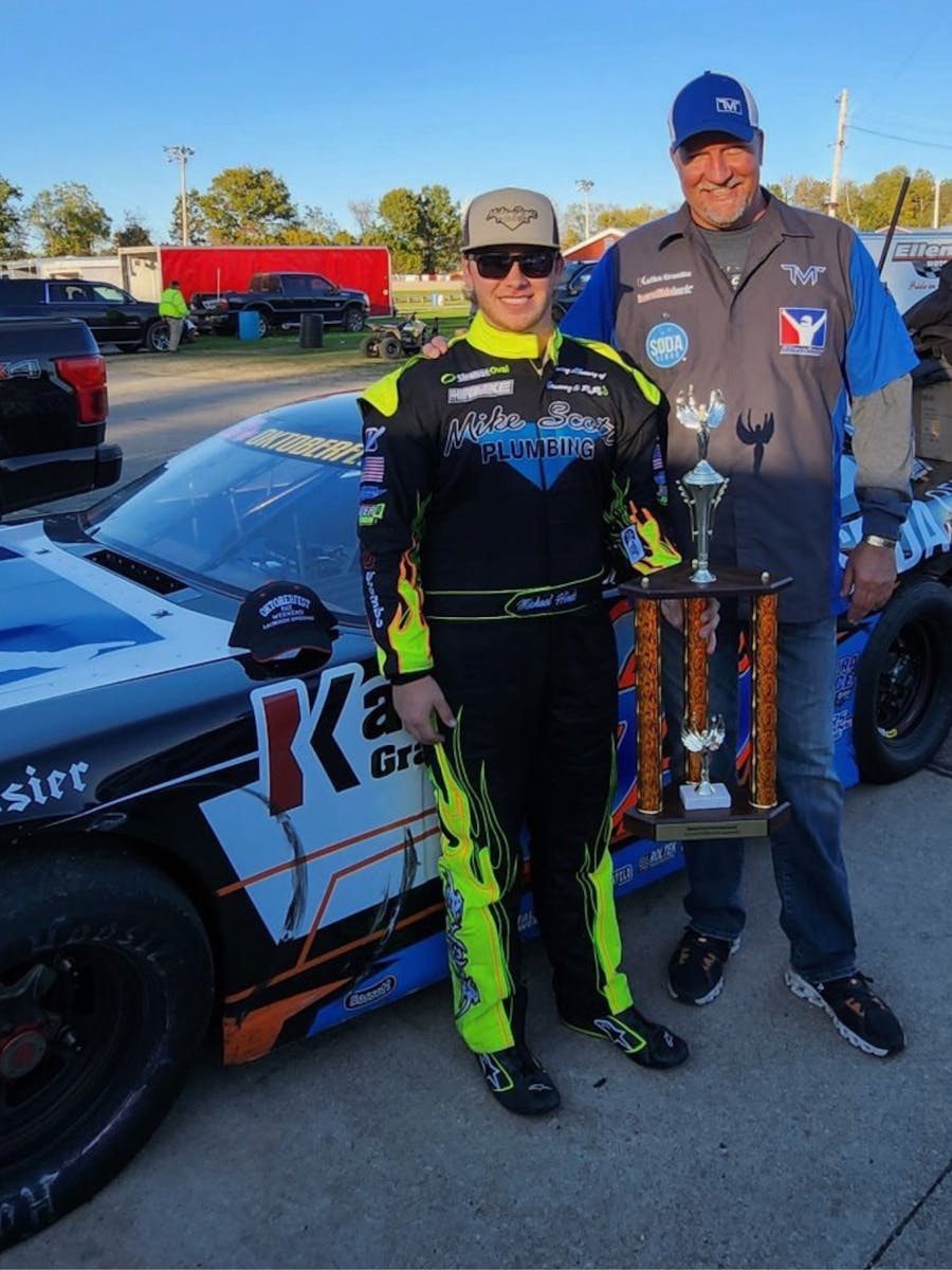 Two people standing in front of a race car, one wearing a neon yellow and black racing suit, the other holding a trophy, at a race track.