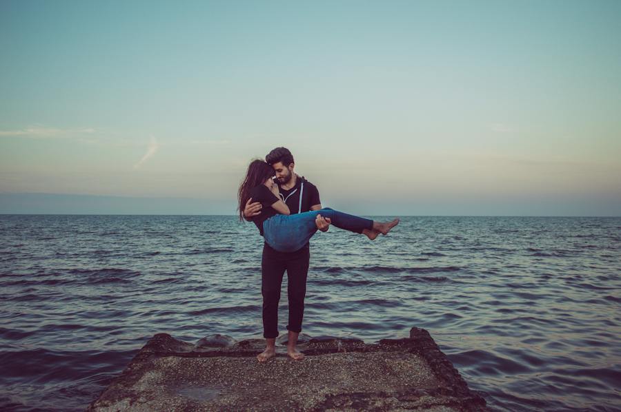 A person is standing barefoot on a concrete platform by the sea, holding another person in their arms. The sky is clear with a hint of clouds, and the water appears calm, reflecting shades of blue and gray as the sun sets on the horizon.