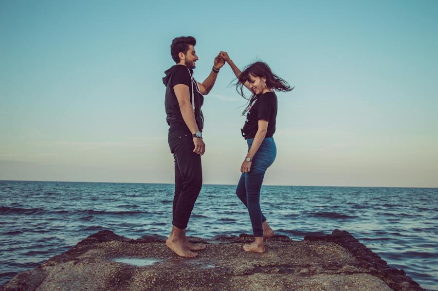 A man and woman dance barefoot at the edge of a rocky outcrop by the sea. The woman twirls under the man's arm, both smiling and enjoying the moment. They are casually dressed against a backdrop of the calm ocean and a clear blue sky.