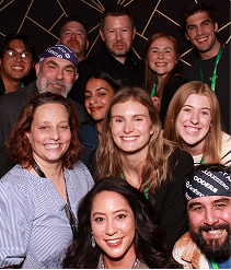 group of 12 people smiling together. 2 are wearing headbands with company logos including Coder, CloudZero, and Grafana